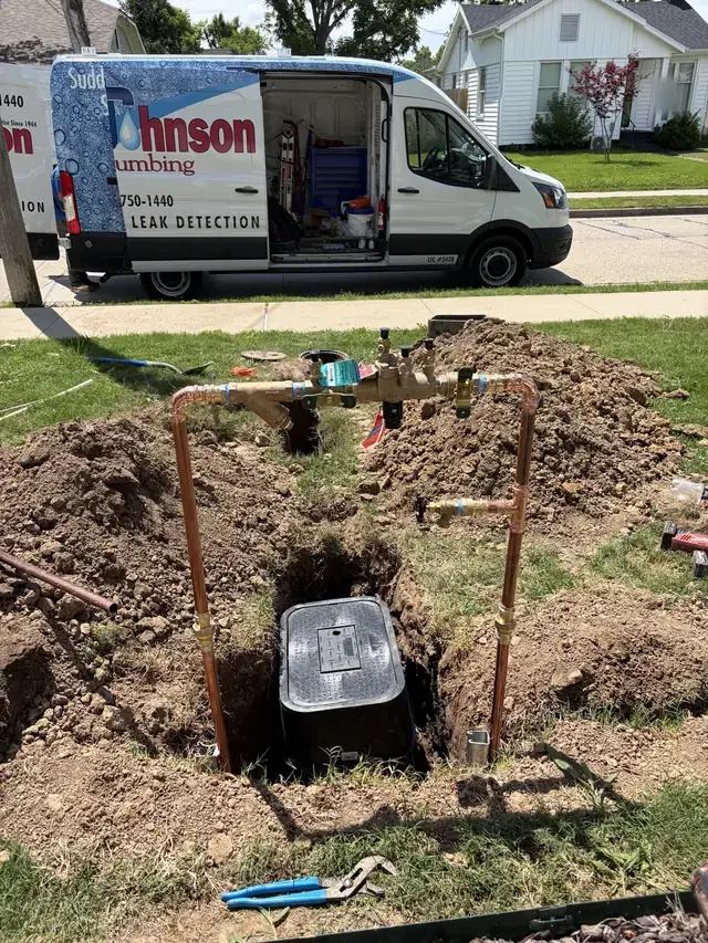 A new backflow assembly on copper risers above a freshly dug meter box in a front yard, the Johnson Plumbing leak detection van parked at the curb