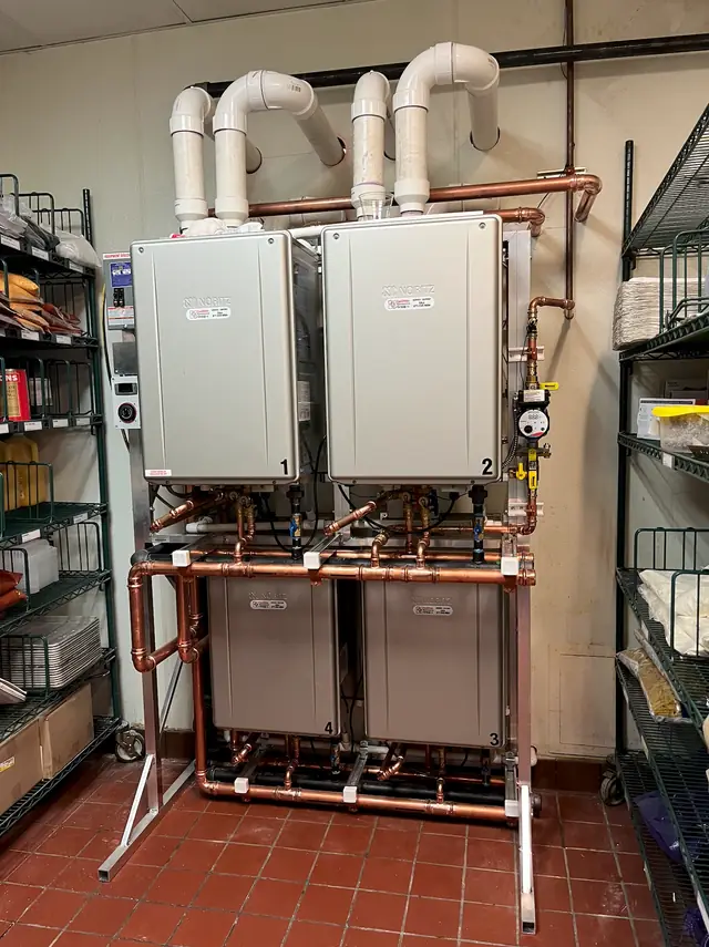 A commercial rack of two tankless water heaters with copper piping and a gas meter, in a storeroom lined with shelving