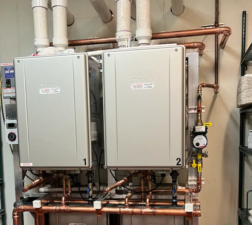 Two wall-mounted tankless water heaters side by side, joined by a copper manifold on a steel rack in a commercial storeroom
