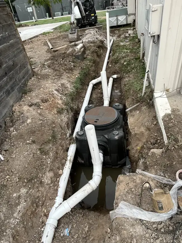 A black grinder basin set in an open trench beside a metal building, white PVC lines tied into it and a mini excavator behind