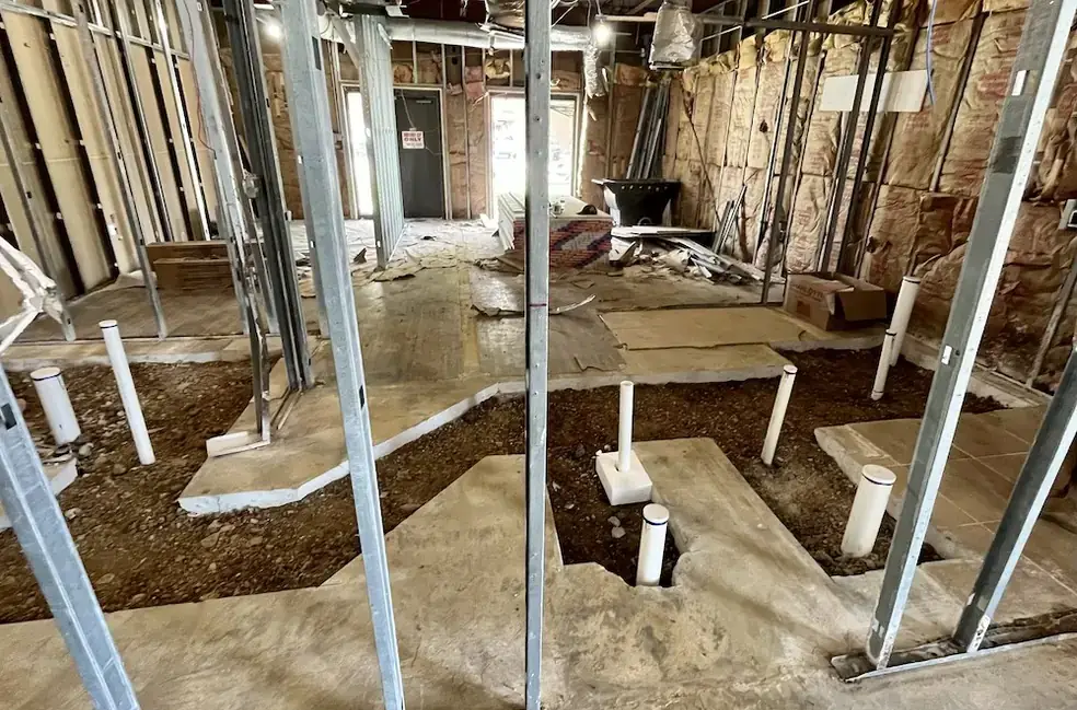 White PVC drain risers standing in trenched gravel between steel wall studs on a commercial rough-in, before the slab is poured
