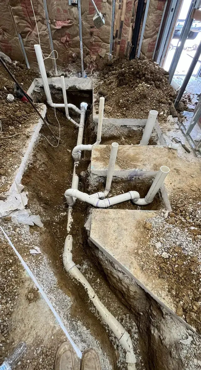 A PVC drain run with wye fittings laid in a trench cut through a concrete floor inside a building under construction