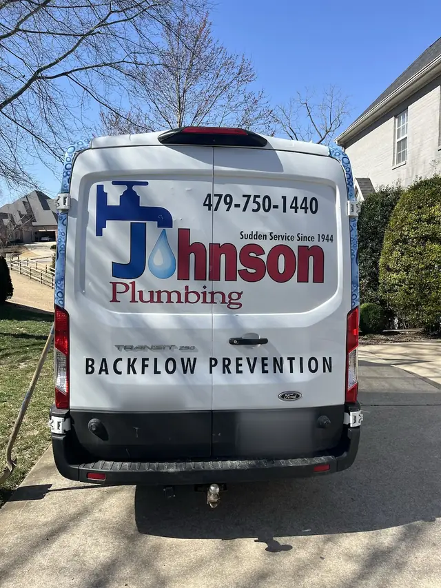 The back of a white Johnson Plumbing van in a residential driveway, lettered 479-750-1440 and Backflow Prevention