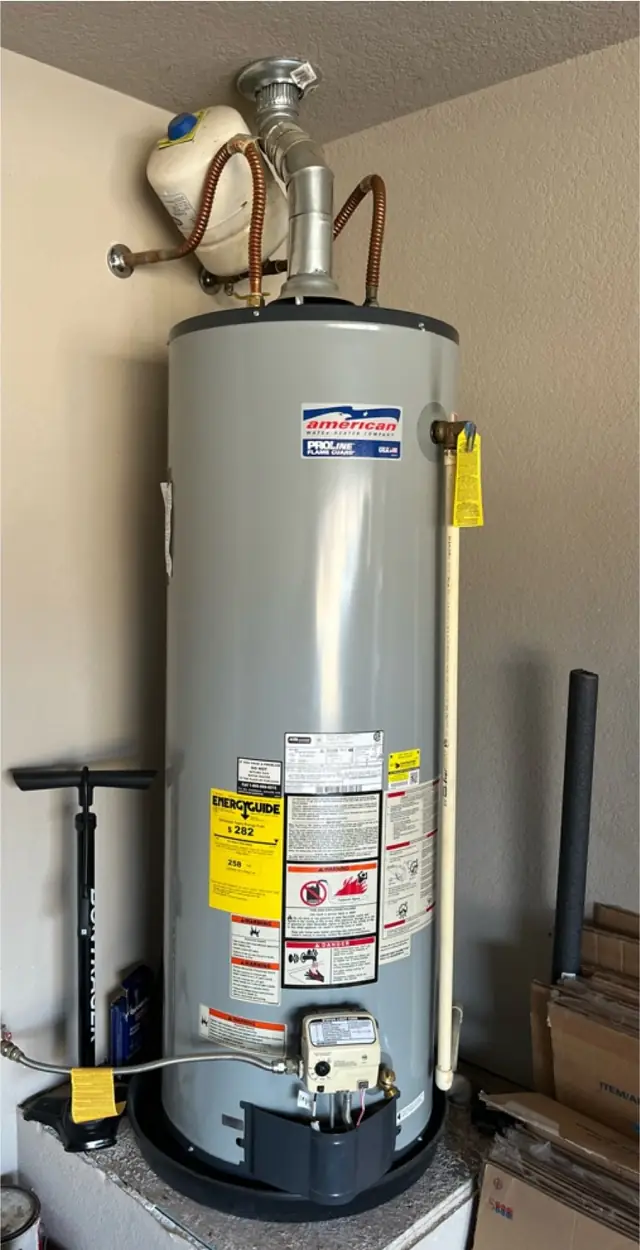 A newly installed gas tank water heater on a drain pan with an expansion tank above it, in a garage corner