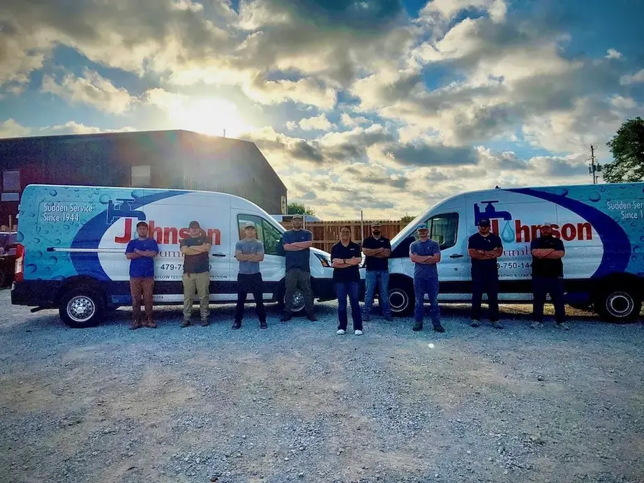 The Johnson Plumbing crew standing with arms crossed between two wrapped vans on a gravel lot at sunset